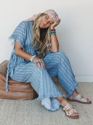A relaxed, dreamy shot of a model seated on a leather ottoman in a chambray striped wide-leg jumpsuit with ruffle trim, styled with stacked turquoise bracelets, a floral headscarf, and neutral sandals — the kind of outfit that looks effortless but wa