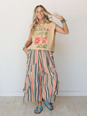 A smiling model in a sandy graphic crop tee and voluminous multicolor striped wide-leg pants styled with turquoise sandals and stacked bracelets — the whole look reads like a summer road trip with good music and zero agenda.
