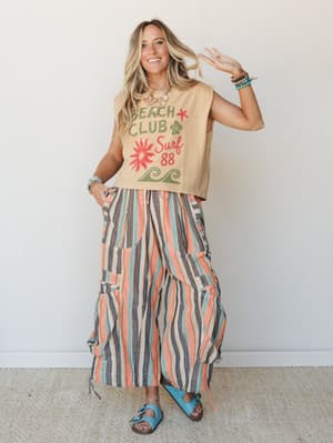 A smiling model in a sandy graphic crop tee and voluminous multicolor striped wide-leg pants styled with turquoise sandals and stacked bracelets — the whole look reads like a summer road trip with good music and zero agenda.
