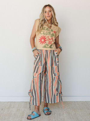 A sun-soaked summer outfit — graphic muscle tee tucked into wide-leg striped crop pants with ruched hem details, styled with turquoise sandals and layered bracelets for a relaxed beach-town energy.