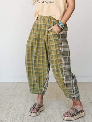 A cropped mid-shot of wide-leg plaid trousers in a moss green colorblocked patchwork, styled with a peach graphic tee, stacked beaded bracelets, and leopard print espadrille sandals — the kind of effortlessly eclectic outfit that makes people stop an