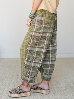 A side-angle shot of relaxed plaid drop-crotch pants in earthy green and tan tones, styled with a soft yellow tee, stacked turquoise bracelets, and leopard print platform sandals — the kind of outfit that has a whole personality.