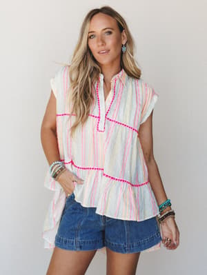 A breezy, color-happy summer look — a tiered sleeveless top in multicolor vertical stripes with hot pink pom-pom trim detail, paired with medium-wash denim shorts and layered beaded bracelets, shot against a soft neutral studio backdrop.