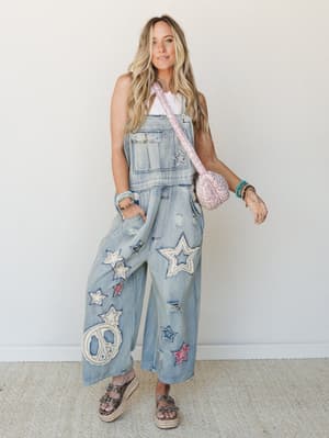 A wide-leg denim overall with star and peace sign appliqués styled over a white tank, paired with a pink quilted crossbody and platform sandals — playful, free-spirited, and very much the outfit you wear when you want people to ask where you got it.