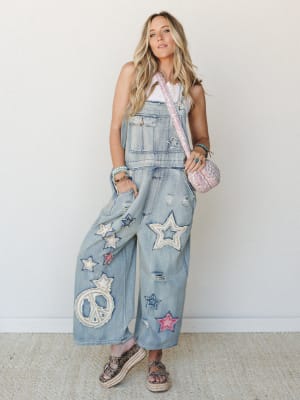 Wide-leg denim overalls covered in star and peace sign appliqué patches styled with a white tank, pink quilted crossbody, and platform espadrille sandals — the kind of outfit that has a whole personality before you even say hello.