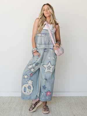 Wide-leg denim overalls covered in star and peace sign appliqué patches styled with a white tank, pink quilted crossbody, and platform espadrille sandals — the kind of outfit that has a whole personality before you even say hello.