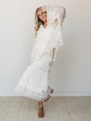 A dreamy ivory tiered ruffle maxi dress with sheer overlay drapes effortlessly on a smiling model in a clean studio setting — romantic, free-spirited, and somehow both bridal and boho at once.