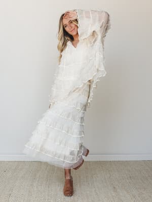 A dreamy ivory tiered ruffle maxi dress with sheer overlay drapes effortlessly on a smiling model in a clean studio setting — romantic, free-spirited, and somehow both bridal and boho at once.