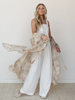 A white wide-leg jumpsuit layered with a flowing floral patchwork kimono caught mid-movement — the kind of outfit that looks equally at home at a bridal shower or a breezy summer market.