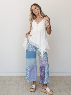 A breezy, free-spirited look pairing a white lace-trim babydoll top with wide-leg patchwork pants in mixed blue florals, paisley, and denim — styled with platform espadrille sandals and layered boho jewelry against a clean neutral wall.