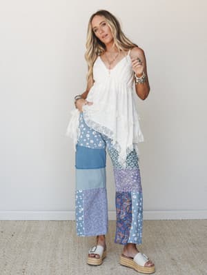 A breezy, free-spirited look pairing a white lace-trim babydoll top with wide-leg patchwork pants in mixed blue florals, paisley, and denim — styled with platform espadrille sandals and layered boho jewelry against a clean neutral wall.