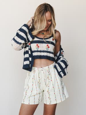 A sun-kissed blonde model smiles softly in a three-piece crochet and embroidered set — striped navy and cream crochet crop top and bell-sleeve cardigan paired with floral embroidered pull-on shorts — shot against a clean light studio backdrop with a