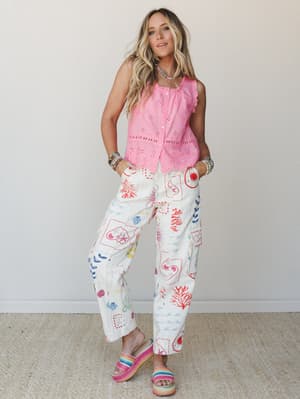 A full-body model shot pairing a bright pink eyelet button-front tank with wide-leg cream pants covered in a playful multicolor ocean-themed print — finished with rainbow platform sandals and stacked bead bracelets for a summer-ready, colorful boho f
