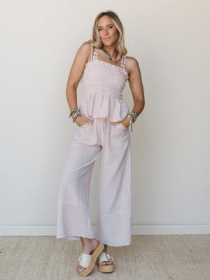A smocked, ruffle-hem top and wide-leg striped pants set in soft blush pink, styled with stacked beaded bracelets, a colorful beaded necklace, and white platform espadrilles — the kind of outfit that works just as hard at brunch as it does at the far
