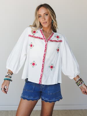 A white embroidered blouse with balloon sleeves and colorful floral cross-stitch detailing is styled with denim scallop-hem shorts — the kind of outfit that feels like a good mood made wearable.