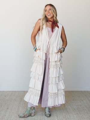 A full-length layered look styled with a cream tiered ruffle duster vest over a soft mauve printed maxi dress, finished with turquoise cowboy boots, stacked bracelets, and a red bandana neck scarf — free-spirited and ready for anything.