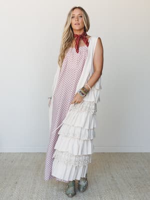 A layered boho two-piece set — a burgundy floral print maxi dress paired with a cream tiered ruffle and eyelet vest — styled with a red bandana, stacked bracelets, and turquoise western boots for a free-spirited, lived-in look.