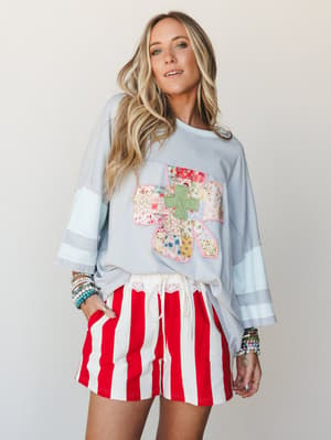 A relaxed, playful two-piece set — an oversized cloud blue sweatshirt with a patchwork floral cross appliqué and striped sleeves, paired with bold red-and-white striped shorts with lace trim, styled with stacked boho bracelets for a free-spirited, li
