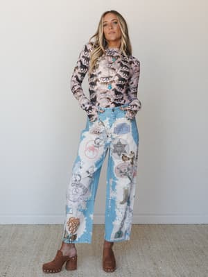A full-body model shot of a playful western-inspired two-piece set — a long-sleeve horse-print mesh top paired with bleach-washed wide-leg jeans covered in cowboy graphic patches — styled with brown platform clogs and a turquoise pendant necklace.