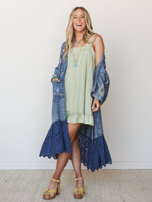 A laughing model layers a sage green ruffle-hem slip dress under a long denim kimono with butterfly embroidery and a navy eyelet ruffle hem — the kind of effortless, mixed-texture outfit that feels like it came together on the best kind of slow morni