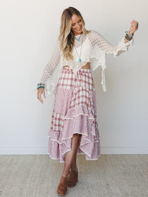 A sun-warmed studio shot of a model dancing in a tiered gingham midi skirt with lace trim and a cropped open-knit shrug — the kind of outfit that makes you want to twirl in a field and then go to brunch.