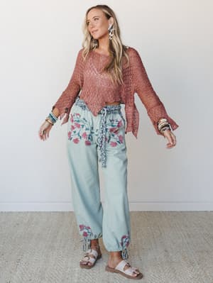 A breezy, expressive outfit pairing a terracotta open-knit crochet crop top with flowy sage wide-leg pants featuring bold floral embroidery — the kind of look that feels like a long weekend somewhere warm.