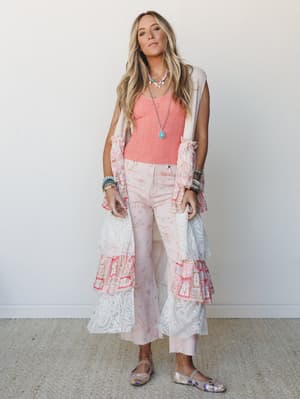 A layered boho look full of soft femininity — a coral lace tank, blush floral flare pants, and a long tiered lace-and-patchwork duster vest styled with turquoise jewelry and metallic flats against a clean white studio wall.
