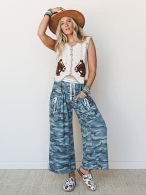 A full-body studio shot of a model wearing a cream crochet horse-motif vest, wide-leg camo pants with drawstring waist, a tan wide-brim felt hat, and cow-print slip-on shoes — the kind of outfit that makes you want to book a road trip immediately.