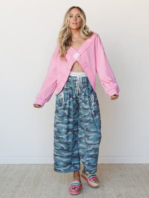 A full-body studio shot of a model in a cropped pink hoodie opened wide and paired with wide-leg camo pants — the kind of unexpected combo that somehow just works, finished with rainbow platform sandals.