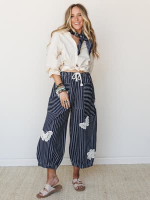 A smiling model in a knotted cream button-down and navy pinstripe wide-leg pants with crochet butterfly and floral appliqués — the kind of outfit that looks effortless but clearly has a whole personality.