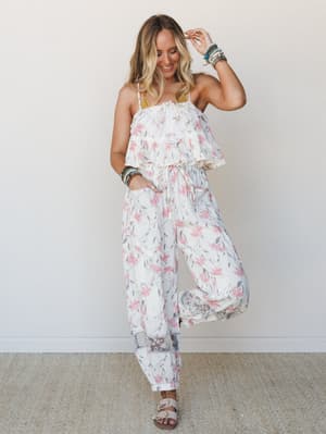 A floral two-piece set styled on a smiling model mid-pose — the ruffled crop top and wide-leg pants feel effortlessly summery, layered with stacked bracelets and flat sandals for an easy, free-spirited look.