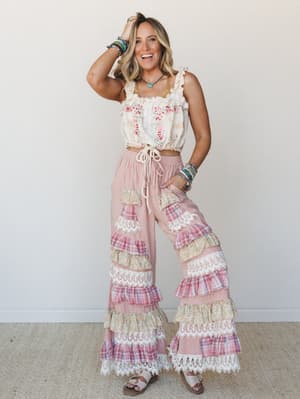 A smiling model in a full boho two-piece set — an embroidered ruffle-strap crop top paired with wide-leg palazzo pants layered in tiered ruffles of pink plaid, floral, and lace trim, styled with stacked beaded bracelets and sandals against a clean ne