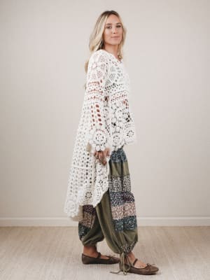 Woman wearing a long, white crocheted cardigan over a patterned dress against a plain background