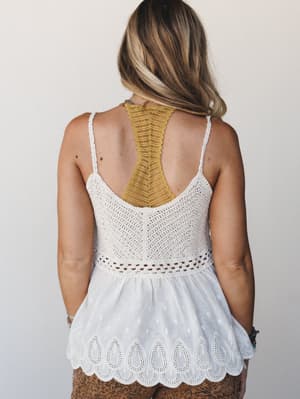 Shot from behind, a model wears a white crochet-knit camisole top with a flowy eyelet hem and scalloped trim — layered over a mustard racerback bralette that peeks through the open back, giving the whole look an effortlessly textured, boho-summer fee