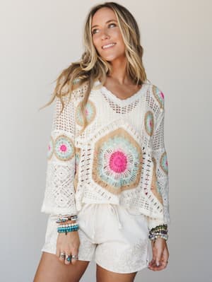 A sun-warmed model smiles off-camera in a cream granny-square crochet top layered over floral-embroidered shorts, stacked with colorful bead bracelets — the whole look feels like a beach town summer morning.