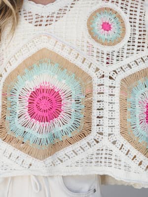 A close-up detail shot of a handcrafted crochet top featuring bold geometric medallions in hot pink, mint, and tan on a cream open-weave base — the kind of piece that looks like it took someone's grandmother three loving weeks to make.