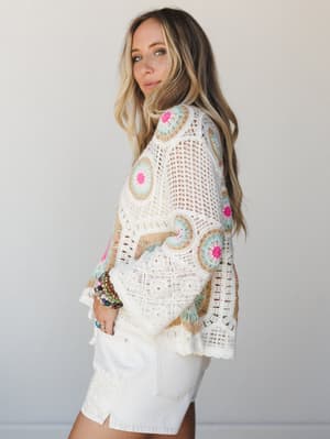 A model shot in soft studio light wearing a relaxed crochet top with colorful granny square motifs — hot pink, sage, and tan against cream — paired with white denim shorts and stacked beaded bracelets, giving the whole look a free-spirited, sunshine-