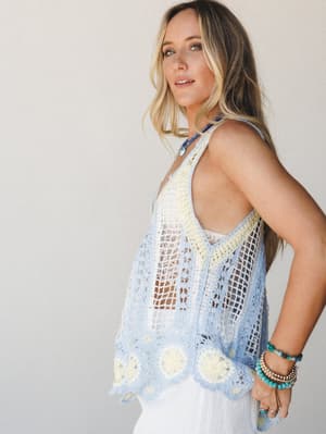 A sun-kissed model in a periwinkle and cream crochet tank layered over a white skirt, styled with turquoise beaded bracelets and a beaded necklace — the kind of outfit that makes a Tuesday feel like a beach town afternoon.