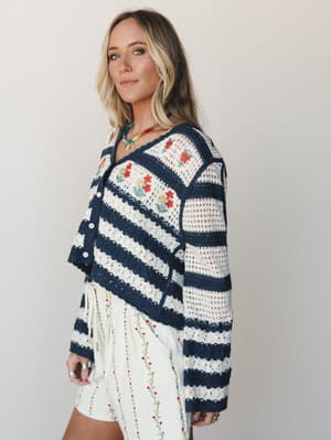A blonde model poses in three-quarter profile wearing a navy and cream striped crochet cardigan with floral embroidery over a cream embroidered mini skirt, styled with a beaded statement necklace — the whole look feels like a garden party and a beach