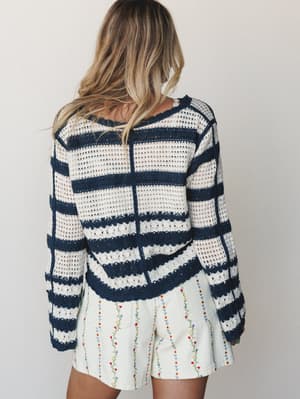 Shot from behind, a model pairs a navy and cream striped crochet cardigan with floral-embroidered cream shorts — the kind of effortless warm-weather outfit that looks like it came together without trying.