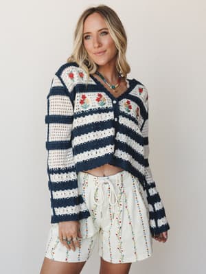 A striped navy and cream crochet cardigan with floral embroidery is paired with ivory embroidered floral shorts, styled with layered beaded necklaces for a playful, cottage-inspired boho look.