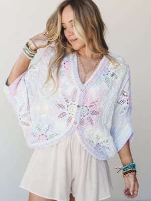 A dreamy pastel crochet shrug in soft rainbow hues worn over a blush mini dress, styled with layered beaded bracelets and a peace sign necklace — the kind of top that looks like it was made from a vintage doily and a good mood.
