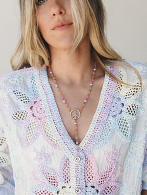 A close-up of a multicolor pastel crochet top with floral and lattice detailing, styled with a beaded peace sign pendant necklace — soft, free-spirited, and very much the kind of thing you'd wear when you want your outfit to say something without hav