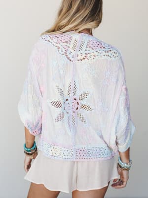 Shot from behind, a sheer white top layered with multicolor pastel crochet appliqué — floral cutwork at the center back, lace trim at the hem and cuffs — paired with a soft blush mini skirt and stacked beaded bracelets for a dreamy, handcrafted feel.