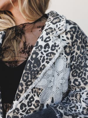 A close-up detail shot of a leopard print denim blazer with raw frayed edges and crochet appliqué detailing, layered over a black lace top — the kind of piece that feels vintage-collected and completely intentional.