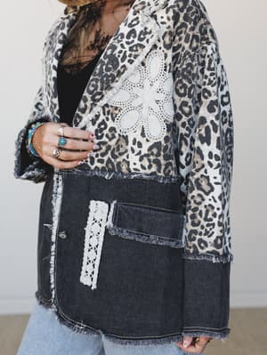 A close-up torso shot of a mixed-media blazer combining leopard print fabric and dark denim panels, detailed with crochet floral appliqué and frayed edges — styled with stacked boho rings and beaded bracelets for a lived-in, expressive feel.