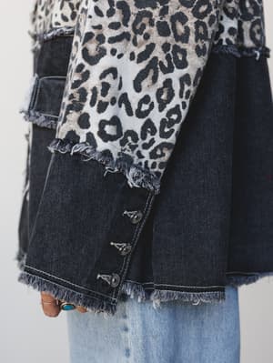 A close-up detail shot of a mixed-media denim blazer with a bold leopard print panel layered over dark washed denim, finished with raw frayed edges and silver concho buttons — worn over light wash jeans with a turquoise ring visible at the hand.