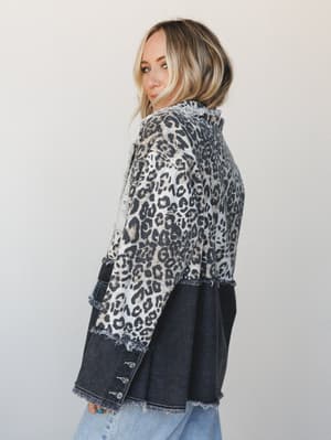 A blonde model glances back over her shoulder showing off the back of a mixed-media blazer — leopard print canvas on top, dark washed denim on the bottom — with raw frayed edges throughout and light blue jeans peeking out below.