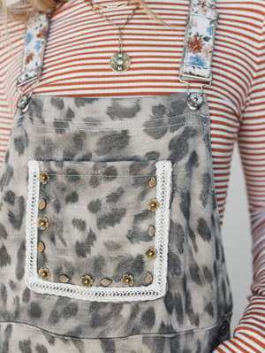 A close-up detail shot of leopard print overalls in a warm tan and grey colorway, styled over a rust striped long-sleeve top — the lace-trimmed pocket with gold floral and leaf studs is the real star here, paired with floral print straps and a delica