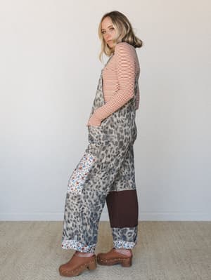 A model shot in profile wearing wide-leg leopard print overalls layered over a rust stripe long-sleeve top, styled with brown wood clogs — the kind of outfit that looks like it was thrown together perfectly without trying.